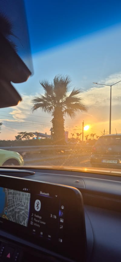 The image shows a view from inside a car, looking out the windshield at sunset. The sky is a gradient of blue to orange, with a bright sun low on the horizon. A palm tree is prominent in the foreground, silhouetted against the sky. There are cars on the road, power lines, and streetlights. The car's dashboard and navigation screen are visible in the lower part of the frame. The navigation screen displays a map and song information, showing "Feelings" by Pascal Junior. The license plate of the car in front reads "TMP 1039". The presence of palm trees, the style of the buildings, and the general ambiance suggest a Mediterranean coastal location, and the specific mention of "Άλιμος, Greece" in the prompt places the scene there.