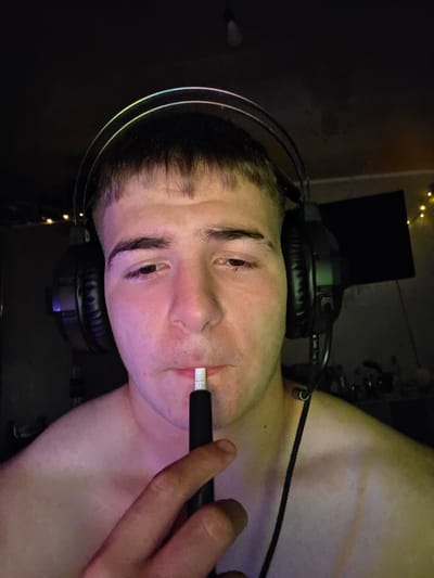 The image shows a close-up of a young person wearing headphones and holding a device to their mouth, possibly a vape or e-cigarette. The background is dark and appears to be an indoor setting with some subtle lighting effects. The person is shirtless, and the lighting casts pink and yellow hues on their skin. The overall impression is one of a casual, possibly gaming-related environment, given the headphones. There is no indication of the image being taken in Aleksandrovo, Bulgaria, or any specific city.