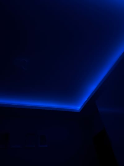 The image depicts a dark room with architectural details illuminated by blue LED strip lighting. The lighting is embedded along the edges of a ceiling recess, casting a cool glow on the surfaces. The overall scene is dimly lit, with the dominant colors being deep blues and blacks. There is a faint, amorphous lighter-colored mark on the upper left portion of the ceiling, which could be a light fixture or a stain. In the lower portion of the image, there are faint outlines of what appear to be framed objects or recessed panels on a wall, also rendered in dark tones. The perspective suggests the photo is taken from a low angle, looking up towards the ceiling. There are no people or animals visible. The location in Vedrare, Bulgaria, is not visually indicated within the image itself.
