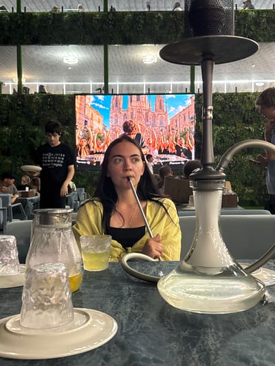 A woman with long dark hair is smoking a hookah in an indoor establishment that appears to be a cafe or lounge. She is wearing a yellow overshirt over a black top and is holding the hookah hose in her mouth. In the foreground, there are several glasses and a pitcher on a dark blue textured table. Behind her, a large screen displays an image of a crowd with their arms raised in front of a building. The walls are decorated with green vertical foliage. A man in a black t-shirt with white text is visible to her left, holding a white plate. Other patrons are dimly visible in the background. The lighting suggests it is evening or the establishment has subdued lighting. The overall atmosphere appears relaxed and social. This scene is set in Derbent, Russia.