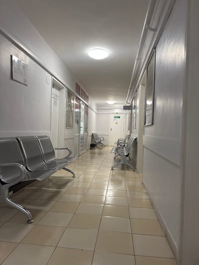The image depicts an interior corridor, likely a waiting area in a public building, possibly a hospital or clinic. The location is in Plovdiv, Bulgaria. The corridor has light-colored tiled flooring and white walls. Rows of metal benches line the corridor on both sides. There are several doors along the left wall, with informational signs and posters. Overhead, there are fluorescent lighting fixtures and various pipes or conduits. The perspective is from one end of the corridor, looking towards the other, and the scene is empty of people. A fire extinguisher is placed by the exit, indicated by the international sign.