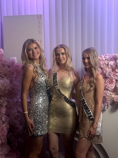 Three blonde women are standing in a row, smiling at the camera. All three women are wearing short sequined dresses and sashes that read "Miss Danmark". The woman on the bottom is wearing a silver sequin dress, the woman in the middle is wearing a gold sequin dress and the woman on the top is wearing a champagne-colored sequin dress. Behind them is a backdrop of white blinds and pink roses. A sign on the left reads "Miss D". Based on the women's sashes, they are contestants in the Miss Danmark competition, and based on the background, they appear to be at the event's location or related activities. While the location is likely within Skovshoved, Denmark, without a clear identifier in the photo, it cannot be definitively located within the city.