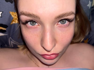 This is a close-up, overhead shot of a young woman's face, specifically focusing on her eyes, nose, and lips. She has light-colored eyes with long, dark eyelashes, and her eyebrows are well-defined. Her skin has a fair complexion with visible freckles across her cheeks and nose. Her lips are full and have a pinkish tint. She is lying down, suggested by the background which appears to be bedding with a patterned fabric, possibly a pillowcase, featuring stars and abstract shapes in shades of blue, yellow, and black. A hint of her shoulder and a delicate gold necklace are visible, indicating she is wearing some clothing. The overall lighting is soft and appears to be artificial, creating a warm glow on her skin. The image does not provide any visual cues or information related to the city of Перепечино, Russia.