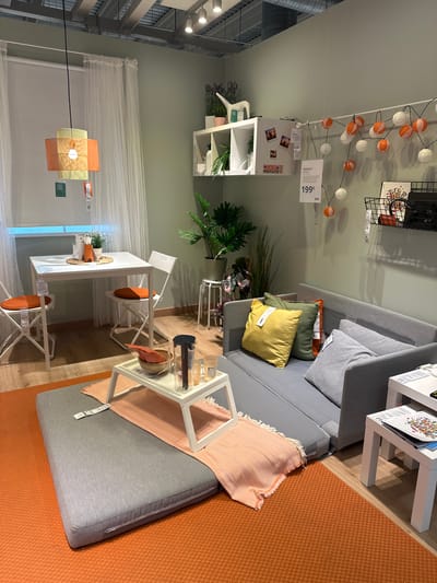 This is a full, eye-level shot of an IKEA showroom in Siero, Spain, featuring a small living and dining area setup. The room is illuminated by artificial lights from the ceiling and a hanging lamp.

In the foreground, a large, flat gray floor cushion is placed on an orange textured rug. To its right, a white side table holds some papers. Behind the cushion is a gray sofa, which appears to be a sofa bed, with several throw pillows in yellow, green, and gray. A white side table is positioned to the right of the sofa.

In the mid-ground, there's a small white dining table with two white folding chairs, each with an orange seat cushion. A white serving tray with a bowl of food, a pitcher, and glasses is placed on the floor in front of the sofa. To the left of the dining table, sheer white curtains hang over a window, and a multi-colored pendant lamp hangs from the ceiling.

The background shows a light green wall with various decorative items. On a shelf are two white square shelves containing plants and decorative objects, including a white watering can. A string of orange and white fairy lights is draped across the wall. A black wire basket hangs on the wall, containing books and other items. Another shelf holds more decorative items.

There are no people visible in the image. The scene is clean and staged, typical of a retail furniture display.