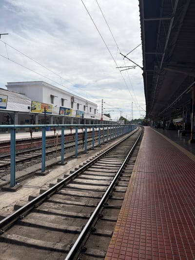 This image captures a railway station platform in Thanjavur, India, with a distinct upward perspective. The scene is dominated by the curved railway tracks leading into the distance, bordered by a red tiled platform on the right and a covered walkway with metal support structures on the left. Overhead wires are visible against a cloudy sky.
A long, white building with multiple windows lines the platform. Several billboards are affixed to its exterior, featuring advertisements and information, some with legible text. One billboard prominently displays "LIC" in yellow and black. Another appears to advertise insurance or financial services with images of people. The architecture of the building is simple and functional, typical of many Indian railway stations.
The time of day appears to be daytime, with diffused sunlight filtering through the clouds. The weather seems overcast. The presence of the railway tracks, platform, and station building clearly indicates the setting is a train station. A few individuals are visible on the platform near the building, but they are small in the frame and not the primary focus. The overall mood of the image is neutral, conveying a sense of everyday transit and the infrastructure supporting it. The perspective emphasizes the expanse of the railway lines and the architectural elements of the station.