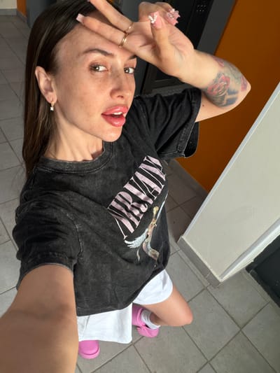 A young woman with long brown hair, wearing a black graphic t-shirt, white shorts, and pink slides, is taking a selfie. She has freckles and is wearing earrings and rings. Her left arm, which is tattooed, is raised to her forehead, and she is sticking her tongue out. The background shows a tiled floor and an orange wall. This scene is not located in the city of Shlisselburg, Russia.