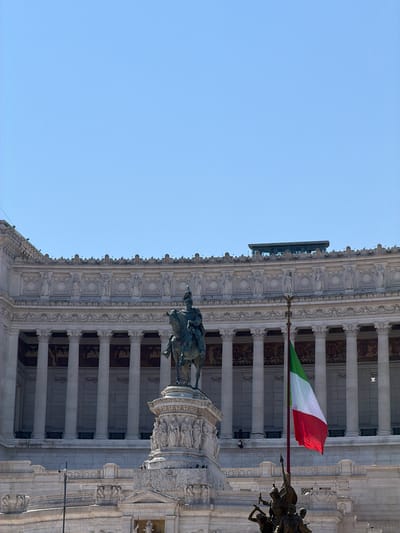 A wide shot captures a monument against a clear blue sky and a building. The monument features a bronze equestrian statue of a man in a military uniform. The statue stands on a tiered pedestal adorned with carved figures. To the right of the statue, an Italian flag flies from a tall pole. The background shows a large, ornate building with classical columns and decorative architectural elements. A few small figures can be seen on the building's facade and in front of it.
