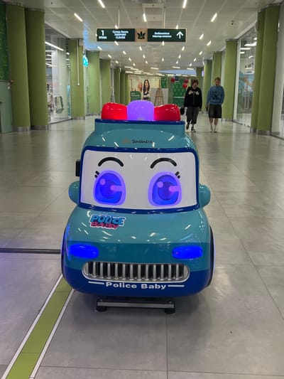 A bright blue and teal police car toy, featuring large cartoon eyes and a red and purple light on top, sits in the foreground of a wide, well-lit indoor walkway. The toy car has "POLICE BABY" written on its front grill. In the background, two young men walk away from the camera. They are wearing casual clothing, one in a black hoodie and dark pants, the other in a blue sweatshirt and shorts. Above them, directional signs are visible on the ceiling, one pointing left and reading "1 ЭТАЖ" (1st Floor) and "Супер Лента DESPORT CLEVER", and another pointing right with an arrow and the text "Библиотека Мастер-классы" (Library Masterclasses). The walkway is lined with large, light green cylindrical pillars and glass storefronts. The floor is tiled in a light gray. The scene appears to be inside a shopping mall or similar public indoor space in Rahya, Russia.