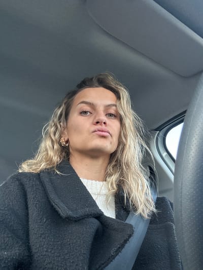 A blonde woman with fair skin and green eyes is pictured from a low angle inside a car. She is wearing a white sweater and a dark coat. A seatbelt is visible across her body. She has on delicate earrings. The car's interior, including the ceiling and dashboard, is visible, along with a glimpse of the outside through the window.