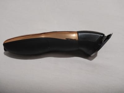 A black and gold electric hair clipper is positioned horizontally on a white, textured fabric surface. The clipper has a black body with a gold accent strip running along its top. The cutting head of the clipper is visible on the right side of the image and appears to be a metallic blade. The fabric surface exhibits subtle wrinkles and a shadow cast by the clipper is visible beneath it. The overall scene suggests an indoor setting, possibly a home, though no specific city, like Moara Domnească, Romania, can be identified from the visual information alone.