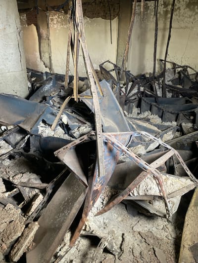 The image shows a scene of devastation and destruction.  The location is the city of Khartoum, Sudan, at coordinates 15.591 latitude, 32.555 longitude.  The environment is characterized by crumbled, broken and twisted metal beams, concrete rubble, and dust.  The scene suggests a collapsed building, potentially the result of an explosion or structural failure.  There are no people present, and the scene is devoid of any human activity or interaction.