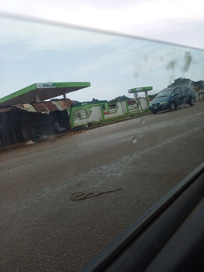 A gas station with a green canopy is visible on the left side of the image. The canopy has the logo "KALAS ENERGY LTD" printed on it. Next to the gas station, there is a makeshift stall constructed with corrugated iron sheets. Some produce, possibly yams or potatoes, are displayed for sale in front of the stall. A dark green van is parked at the gas station on the right side of the image. The road in front of the gas station appears wet, suggesting recent rain. A coiled rope is lying on the road in the foreground. The sky is overcast. The scene is likely located in Panyam, Nigeria, as indicated by the context.