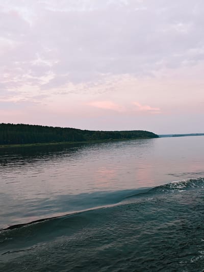 A wide shot shows a tranquil body of water with a dark green treeline stretching across the horizon. The sky above is a pale lavender with streaks of pink clouds. Gentle waves ripple across the water's surface, catching the soft light. In the foreground, a small wave breaks, its white foam contrasting with the darker water. The scene is serene and quiet. It's a dusk view of the Svir River in the city of Nakhval'skoe, Russia.