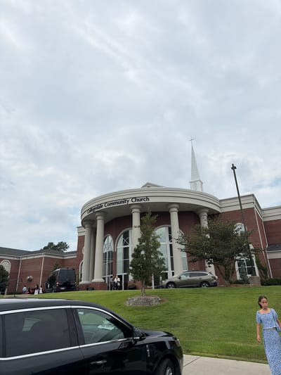 The image shows the Collegedale Community Church in Collegedale, Tennessee, United States of America. The church has a red brick facade with large arched windows and white columns. A steeple with a cross is visible at the top of the building. Several people are seen entering and exiting the church, and a black van is parked in front. A young girl in a blue floral dress walks on the right side of the frame, and a black car is parked in the foreground. The sky is overcast with clouds.