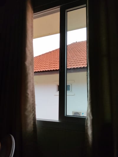 This media file is a photograph taken from inside a room, looking out through a window. The foreground is dominated by dark, thick curtains on either side of the frame, creating a sense of looking through a veil. The window itself is a sliding glass door, partially open, revealing a view of a neighboring house.
The house visible outside has a distinctive red-tiled roof, typical of many houses in tropical regions. The walls of the house are painted white. Two windows are visible on the exterior wall, one small and square, and another larger one showing an air conditioning unit mounted on the exterior. The air conditioning unit appears to be a Daikin brand, as indicated by visible text and logo on the unit.
The sky above the house is bright and appears to be a clear day, suggesting it is daytime. Given the location context of Ban Patan, Thailand, this likely means it's warm or hot outside. The angle of the light suggests it could be mid-morning or afternoon. The image evokes a sense of quiet observation, perhaps from someone indoors looking out at their surroundings. There is no clear indication of any people or specific activities taking place, other than the general scene of a residential area. The overall mood is peaceful and somewhat secluded, due to the framing by the curtains.