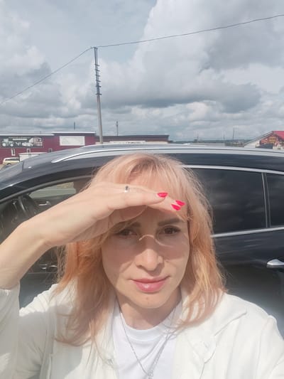 A medium shot photograph captures a woman from the chest up, standing in front of a black SUV. She has peachy-blonde hair with bangs and is wearing a white top and jacket. Her right hand is raised to her forehead, shielding her eyes from the sun. She is wearing a ring on her ring finger and bright pink nail polish. Her lips are slightly parted, and she is looking directly at the camera. The background shows a cloudy sky, a utility pole with a ladder, and buildings with signage. The overall impression is a candid selfie taken outdoors. The location is identified as Belka, Russia.