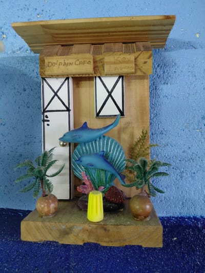 A wooden miniature diorama of the "Dolphin Cafe, Key Largo" is positioned on a blue surface. The diorama features two stylized blue dolphins, miniature palm trees, faux coral, and small wooden "windows" marked with black "X"s. The wooden frame displays "Dolphin Cafe" and "Key Largo" inscribed on its side. A yellow object is visible near the top right corner of the diorama. The background shows a textured blue surface. It is not possible to confirm the diorama's location in Cutler Bay based on visual information.