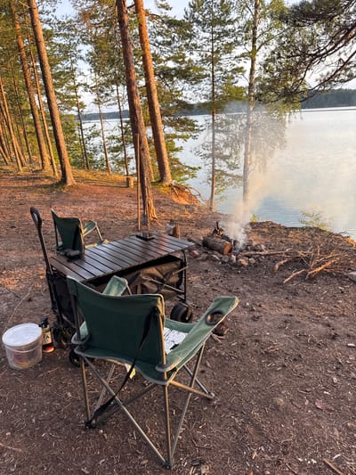 A campsite is set up on a sandy and wooded shore of a lake at sunset. The scene features a portable table with two green camping chairs, one of which is in the foreground. Behind the table, a wagon is visible, along with a portable grill. A campfire burns on a rock-lined pit to the right of the table, with smoke rising into the air. Various pieces of firewood and branches are scattered around the campfire. A clear plastic container and a can of compressed gas are on the ground near the wagon. The background shows a calm lake with trees on the opposite shore. The ground is covered with pine needles and fallen branches. The location is implied to be in Kutuzovskoe, Russia, based on the provided context.