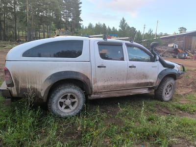 The image shows a silver Toyota Hilux pickup truck covered in mud, parked on a dirt and grassy area. The truck has a white canopy over the bed and a snorkel attached to the driver's side. In the background, there are trees, power lines, a wooden building, and some construction equipment. The scene appears to be in a rural or semi-rural area, possibly on the outskirts of a settlement. The lighting suggests it is late afternoon or early evening. The location is identified as Гремучий, Russia.