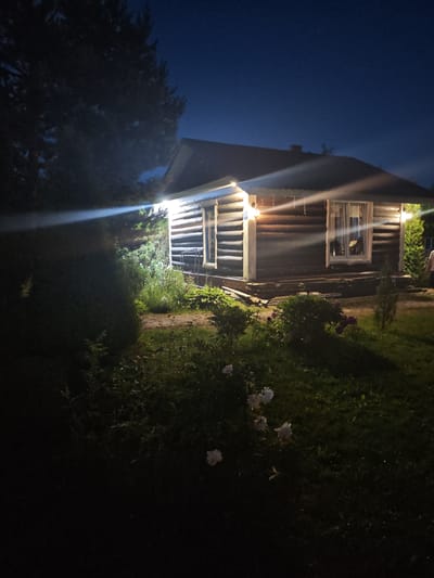 A wooden cabin is illuminated by exterior lights at night. The cabin is dark wood and has a sloping roof with lights strung along the eaves. There is a window on the front of the cabin with curtains. In front of the cabin is a grassy yard with some bushes and flowers. A tall tree is to the left of the cabin, and the sky is dark blue, indicating nighttime. The location is identified as Надежда Снт, Russia.