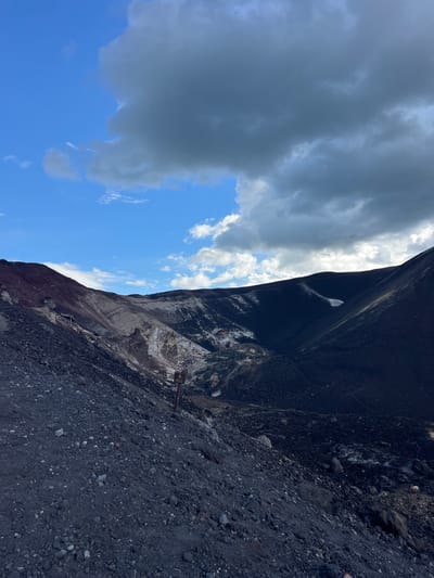 The image depicts a steep, rocky slope with a clear blue sky and dramatic, dark clouds above. The terrain is composed of dark, granular volcanic rock and larger stones, with areas of lighter-colored, possibly mineral-rich deposits visible on the slope. A small yellow excavator is parked on the slope, suggesting ongoing excavation or maintenance work. A wooden signpost with a yellow warning symbol is also present. The light suggests it is daytime, and the presence of clouds indicates varied weather conditions. The scene has a rugged, natural feel, possibly a volcanic crater or a quarry. There is no visible text on the sign other than the symbol.