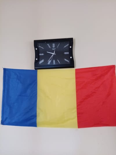 A close-up, eye-level, medium shot captures a rectangular digital clock mounted on a plain, light-colored wall. Below the clock, a Romanian flag is draped horizontally across the bottom half of the frame. The flag is divided into three equal vertical stripes: blue on the left, yellow in the middle, and red on the right. The clock has a black frame and a black face with white Roman numerals. The hour and minute hands are white. The scene is lit from the front, casting no shadows. The overall impression is one of domestic simplicity, with no indication of any specific location within the city of Saurimo, Angola.
