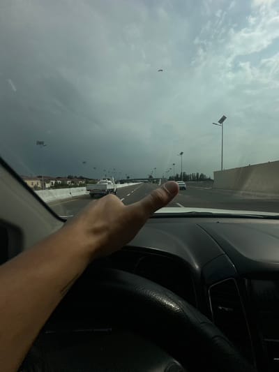 The image shows a view from inside a car, looking out through the windshield. The perspective is from the passenger side. A person's thumb is prominently visible in the foreground, pointing towards the road ahead. The road stretches into the distance and appears to be a highway with multiple lanes. There are several vehicles on the road, including trucks and cars, and traffic seems to be moving.

The setting is an outdoor environment with a cloudy sky. The clouds are dense and gray, suggesting overcast weather, possibly with rain imminent or recently passed. There are streetlights or lampposts lining the road at intervals. In the middle ground, there are some buildings and trees visible, with more trees lining the far horizon. A tall, light-colored wall or barrier runs along the side of the road.

The time of day appears to be daytime, given the ambient light, but the overcast sky makes it difficult to determine the exact time. The mood seems neutral, with the focus on the journey and the road ahead. There is a small, dark object visible in the sky, possibly a bird or a kite. No visible text is present in the image. The provided location context is Jaukаshty, Uzbekistan, which might suggest the architectural style of the buildings or the type of vegetation, though these are not distinctly identifiable from the image alone.