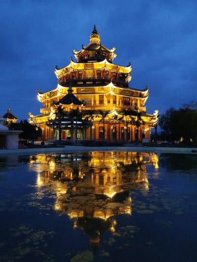 A five-tiered pagoda building is illuminated in yellow light in the city of Phú An, Vietnam, reflecting in a body of water below. The sky is a deep blue. Trees are to the right of the pagoda, and another building is to the left. Algae or other vegetation is visible in the water.