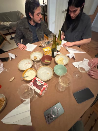 The image depicts an indoor social gathering at a private residence or similar venue in Roanne, France. The scene centers on a large, round table covered with a brown fabric, illuminated by overhead artificial lighting.

Two individuals are prominently visible at the table. On the left, a male with dark hair and a beard, wearing glasses on his head and a dark patterned jacket over a dark shirt, rests his left hand on a piece of white paper while looking towards the right. On the right, a female with long dark hair, wearing a dark short-sleeved top, is actively folding a piece of white paper with both hands, looking down at her task. Additional hands and arms indicate at least one more person is seated at the table, out of frame to the lower right.

The table is occupied by numerous items, suggesting recent food and drink consumption and a shared activity. These include two mostly empty green glass wine bottles, several empty or partially full wine glasses containing residual white wine, and a wine cork and corkscrew. Multiple ceramic bowls of varying sizes and colors contain remnants of snacks, such as crumbs, a single potato chip, a cucumber slice, and a white dip. A small cardboard box of "LU" branded crackers, a plastic container with clear dividers, and a red-and-white cardboard box labeled "Azura" (featuring a strawberry graphic) are also on the table. A bowl of peanuts is visible in the lower left corner. Several pieces of white paper, some folded into paper airplanes and others flat, are scattered across the table, consistent with the activity engaged in by the visible individuals. Two smartphones, one with a dark MagSafe-compatible case and the other with a dark screen, are placed on the table surface. The background shows a dimly lit room with a light-colored wall, a sofa, and a bookshelf.