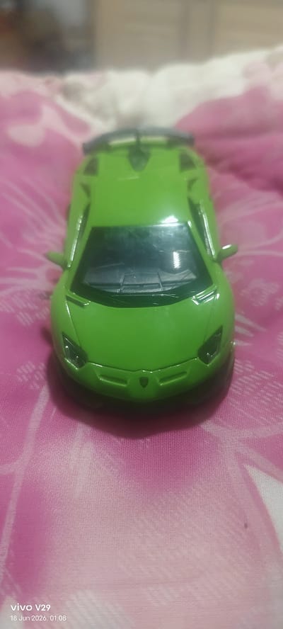 A green toy sports car is displayed from a high angle, positioned centrally on a pink and white patterned fabric. The car is in focus, while the background is blurred, creating a shallow depth of field. The fabric appears to be a blanket or sheet with a soft texture. The timestamp "vivo V29 18 Jun 2026, 01:08" is visible in the bottom left corner of the image. There are no people present in the image, and no specific actions or interactions are depicted. The scene is static and takes place indoors, as indicated by the fabric surface and the lighting. There is no information available regarding the city of Pulodolang, Indonesia.