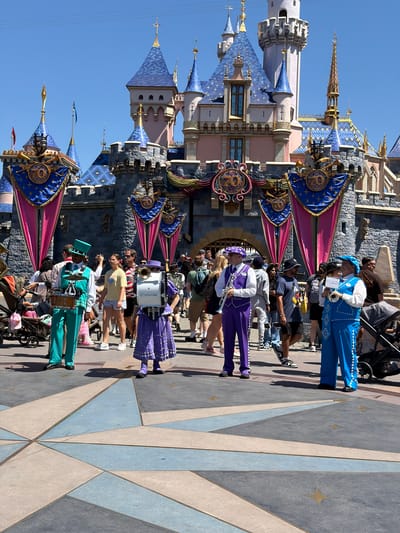 This image captures a bright, sunny day at Disneyland in Anaheim, California. In the foreground, a group of performers dressed in colorful marching band uniforms stand on a patterned tiled plaza. One performer in a teal suit plays a drum set, while others in purple and blue outfits play brass instruments. Behind them, the iconic Sleeping Beauty Castle is visible, adorned with festive banners celebrating the "70"th anniversary. A crowd of park-goers, including families and children, are seen walking and observing the performers. Strollers and shopping bags are also present, indicating a typical theme park environment. The overall atmosphere appears lively and celebratory.