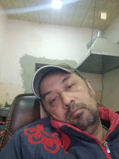 A man with a salt-and-pepper beard and a gray baseball cap tilted to the side is angled toward the camera. He wears a dark blue jacket with red trim and an ornate red design on the shoulder. A ceiling-mounted fixture with a chain and a bright light occupy the background wall. The man rests in a chair with visible woven details. The background wall shows some patches and imperfections in the paint.