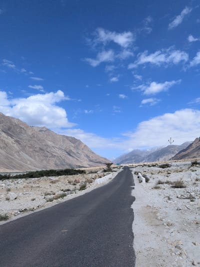 This is a photograph of a desolate road in Sasoma, India, under a bright blue sky with scattered white clouds. The road itself is asphalt and curves into the distance, flanked by a dry, rocky landscape. On the left side of the road, a rugged, barren mountain range rises sharply, its slopes showing layers of rock and sparse vegetation. On the right side of the road, the terrain is flat and covered with pebbles and rocks, with a few hardy bushes. In the distance, more mountains are visible, some with snow-capped peaks. Several utility poles with wires are scattered along the right side of the road, indicating some form of infrastructure. The overall scene is one of vastness and isolation. There are no people or animals visible in the image.