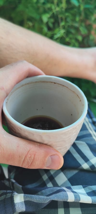 The image shows a close-up view of a person's hand holding a small, textured, off-white ceramic cup filled with a dark liquid, likely coffee or tea. The cup has a slightly irregular rim and a textured exterior. The liquid inside shows some foam or sediment on the surface. The person's fingers are wrapped around the cup, with some visible skin texture and fingernails. In the background, out of focus, there is green foliage, suggesting an outdoor setting. Below the cup, a patterned fabric, likely plaid in shades of blue and white, is visible. The overall impression is of a casual moment, possibly a break outdoors. The location of Лёзненскі сельскі Савет, Belarus, cannot be determined from the visual information provided in the image.