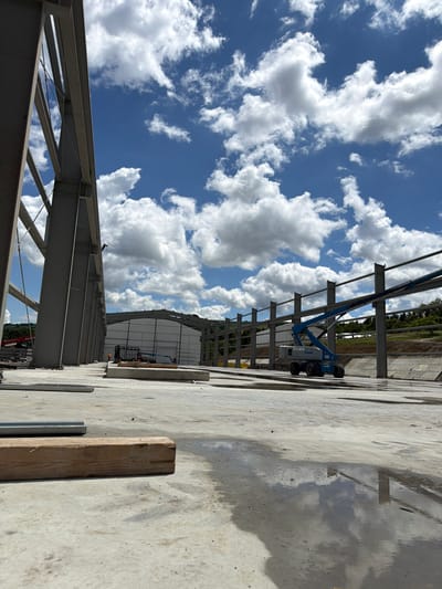 The image shows a construction site in Skrzydlna, Poland, under a blue sky with scattered white clouds. In the foreground, a large piece of lumber lies on a wet concrete surface. Beyond this, a construction project is underway with exposed steel beams forming the framework of a building. A blue Genie lift is positioned near the beams, and in the background, a partially constructed building with white siding is visible. The overall scene depicts an active construction environment.