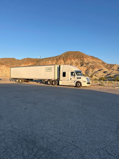 A white semi-trailer truck is parked on an asphalt surface. The truck is mostly visible from its side, with the trailer extending to the left and the tractor to the right. The trailer is long and white with "Premier Trailer Leasing" logo on the side. The tractor is white and has "I GO HAUL LLC" written on the door. The truck is parked in front of a landscape with arid hills and sparse vegetation. The sky is clear and blue. The image appears to be taken in a rural or desert area, possibly a rest stop or parking lot for trucks. The location is not Salina, Kansas, as the landscape does not match that region.