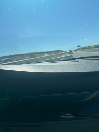 The image shows a view from inside a car driving on a highway. The sky is blue and clear with some specks of dust on the windshield. The highway has multiple lanes and other cars are visible in the distance. There are trees and hills on the side of the road. The dashboard of the car is visible in the foreground, and there is a reflection of a person's hand holding a phone on the dashboard. The scene is consistent with driving on a sunny day. Without further information, the exact location within Vratitsa, Bulgaria, cannot be determined from the image alone.