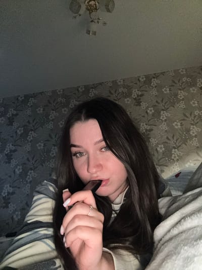 The image, taken indoors and likely in a residential setting in Osentsy, Russia, shows a young woman with dark hair and fair skin. She is positioned in a close-up shot, with her face and upper torso filling most of the frame. The woman is wearing a sweater with a striped pattern. Her right hand is raised towards her mouth, holding a dark, rectangular object, possibly a piece of chocolate or a small electronic device. She appears to be in the process of bringing the object to her lips. Her gaze is directed slightly off-camera, and she wears a delicate ring on her right index finger. The background consists of patterned wallpaper, suggesting a bedroom or living area. A light fixture is visible on the ceiling in the upper portion of the image. The lighting is dim, creating a soft, intimate atmosphere.