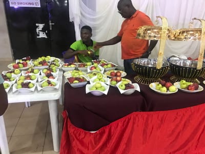 Inside a room in Ife, Nigeria, there is a fruit display on a white table and a table covered with a maroon and red cloth. On the white table are several white bowls filled with green apples, red apples, and grapes. A man in a green shirt is seated at the white table and appears to be handling an apple. Another man in an orange shirt stands next to the table covered with a maroon and red cloth and appears to be extending his hand. On the maroon and red table are more bowls of apples and grapes, as well as two large serving dishes with gold covers and handles. In the background, there is a white curtain, a dark door with a "No Smoking" sign, and light-colored walls.