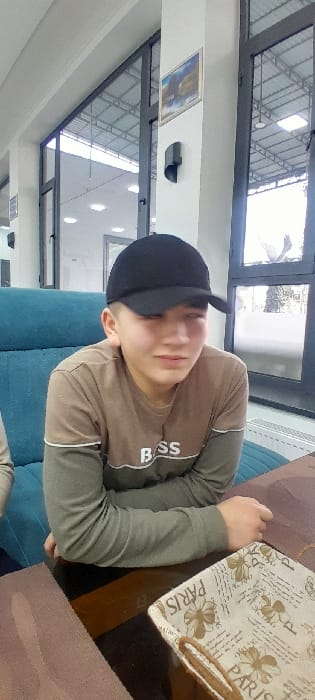 A young male is seated at a dark brown, reflective table in what appears to be a cafe or restaurant located in Chinabad, Uzbekistan. He wears a black baseball cap and a two-toned long-sleeved shirt: light brown on top with "BOSS" printed in white, and dark grey or olive green sleeves. His eyes are partially closed or squinting, and his mouth is slightly downturned, suggesting a grimace or discomfort. His hands rest on the table. To his left, part of another person's light-colored clothing is visible. On the table to his right, a light-colored fabric basket with "PARIS" text and a floral pattern is present, next to a dark brown placemat. Behind the male is a teal or turquoise upholstered bench. The background features large, dark-framed windows that reveal an outdoor structure with a metallic roof and foliage. A white pillar separates window sections, displaying a framed picture with blue and white tones and a black wall-mounted light fixture. The ceiling is white with recessed lighting.