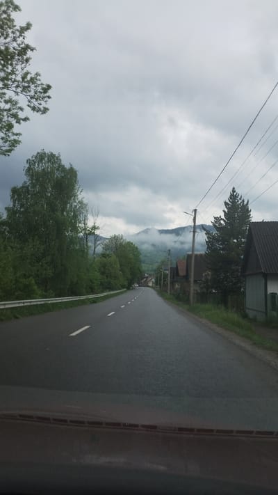 The image shows a view of a wet asphalt road in Yasinia, Ukraine, with a mountain obscured by clouds and fog in the background. Power lines stretch across the sky above the road, and buildings are visible along the road's edge. Trees and vegetation are present on the sides of the road. The image appears to be taken from inside a vehicle.