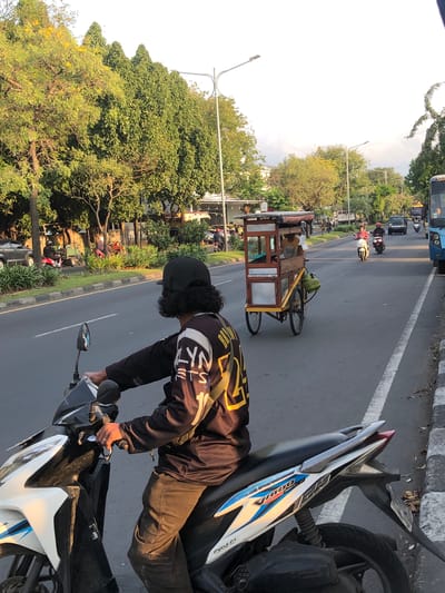 In the city of Gelam, East Java, a man wearing a black baseball cap and a black long-sleeved shirt with yellow and white lettering rides a white and black motorcycle. He is positioned in the foreground, facing away from the camera and looking towards the right side of the frame. His right hand is on the handlebar. To his right, a food cart with a wooden frame and a canopy is being pushed by a person partially visible inside. The food cart has two wheels. The scene unfolds on a paved road with a white lane marker. In the background, several trees line the left side of the road, and more trees are visible on the right, interspersed with buildings. Other vehicles, including motorcycles and cars, are present on the road. The sky is bright, suggesting daylight.