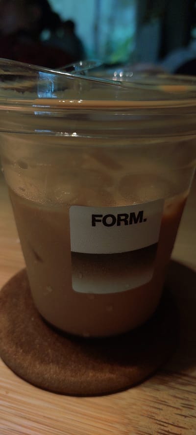 The image shows a close-up of a clear plastic cup filled with a light brown iced beverage, likely coffee or tea with milk, resting on a circular brown coaster. The cup has a white label with the word "FORM." printed in black, sans-serif font. Condensation is visible on the outside of the cup, indicating it is cold. The background is blurred, showing indistinct dark shapes and some blue and green hues, suggesting an indoor environment with ambient lighting. The cup is placed on a wooden surface, possibly a table. There is no indication of a specific location within Sampora, Indonesia, or any people present.