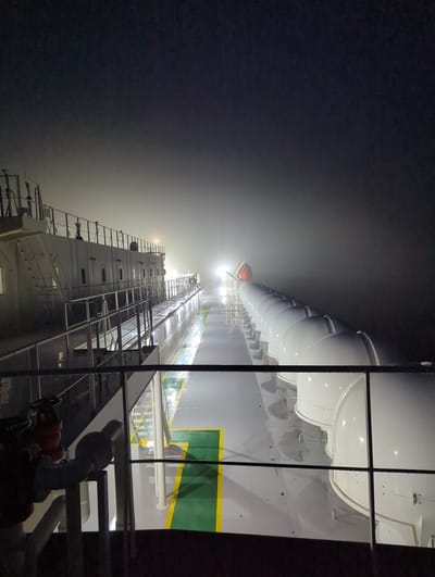 This is a photograph taken from the perspective of being on a ship at night. The image is dominated by the structure of the ship, with lifeboats visible along the side. The scene is illuminated by artificial lights from the ship, casting a bright glow that pierces through a thick fog or mist. The fog obscures the surrounding environment, creating a sense of isolation and mystery. The perspective is angled, looking upwards along the length of the ship, highlighting the towering structure and the numerous lifeboats. The water is not visible, but the presence of the ship and the fog suggests a maritime setting. There is no visible text in the image. The time of day is clearly night, due to the artificial lighting and the darkness of the sky, which is further enhanced by the fog. The weather condition is foggy, creating a dramatic and somewhat eerie atmosphere. The location cues suggest a large vessel, possibly a ferry or a cargo ship, operating in open water. The image evokes a sense of solitude and perhaps a hint of suspense due to the limited visibility.