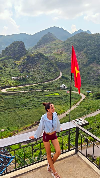 A woman is standing on a balcony overlooking a mountainous landscape in Lũng Phìn Commune, Vietnam. She is wearing a light blue jacket, red shorts, and pink sandals. Her hair is tied up in a bun. She is leaning on the balcony railing, looking off into the distance. A Vietnamese flag is positioned on a pole nearby. The background features rolling hills covered in green vegetation, a winding road, and scattered houses. The sky is partly cloudy.