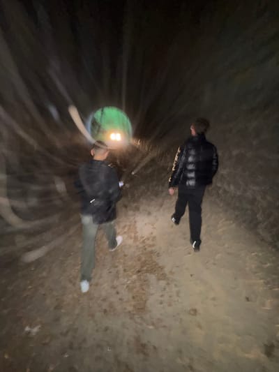The image provides an overhead view of two individuals exploring a dark, enclosed tunnel or large pipe in Bures-sur-Yvette, France. The primary light source is a bright, green-yellow headlamp worn by the person in the foreground, creating significant lens flare and a radial blur effect that suggests movement or a long exposure.
The individual further away is dressed in a plaid shirt over a dark garment, dark pants, and white-soled shoes. The person closer to the camera wears a dark jacket or sweatshirt, light-colored pants, and white shoes, and appears to be carrying a dark bag or backpack. Both figures are oriented as if moving forward through the space.
The environment is dim, with dark, seemingly damp, curved walls and a textured, earthy or concrete floor. The complete reliance on artificial light underscores the subterranean or deeply enclosed nature of the location. No specific text is visible within the image, and the subjects' expressions are not discernible. The overall impression is of an exploratory activity within a confined and dimly lit passage.
