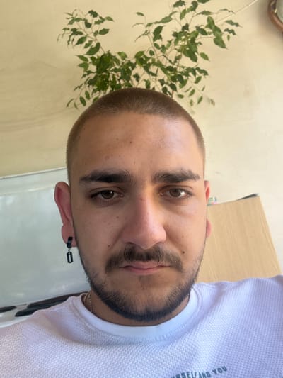 A close-up portrait, rotated ninety degrees counter-clockwise, depicts a young adult man looking directly at the camera with a calm and neutral expression. He has a very short, buzzed haircut in a light brown color, complemented by thick, dark eyebrows and dark brown eyes. A short beard or prominent stubble frames his jawline and chin. He wears a white, short-sleeved top made of a textured, pique knit fabric. A silver chain is visible around his neck, and a distinctive black, rectangular accessory, possibly an earpiece or an earring, hangs from his left ear.
The background behind him is split. To his left, there is a plain, light yellowish-beige wall, with a vibrant green, leafy plant (possibly a ficus) positioned behind his head and shoulder. To his right, the background is a lighter, almost white surface, with a glimpse of a light wooden or neutral-toned structure.
The lighting is bright and even, suggesting a daytime setting, either indoors or in a well-lit shaded area. The man is the clear focus of the image, appearing to be posing for the photo. A partial phrase is visible on his white shirt, which, when properly oriented and read, appears to be "YOURSELF AND YOU" (likely part of a larger motivational phrase). This scene is situated in Staro Selo, Bulgaria.