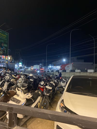 At night in Seuneubok Teungoh, Indonesia, a densely packed row of motorbikes lines a street. A white car is parked to the right, partially obscuring the view. Utility wires crisscross overhead. The scene is illuminated by streetlights and the headlights of distant traffic. A billboard featuring the text "R Syariah Bank Aceh" is visible on the left.