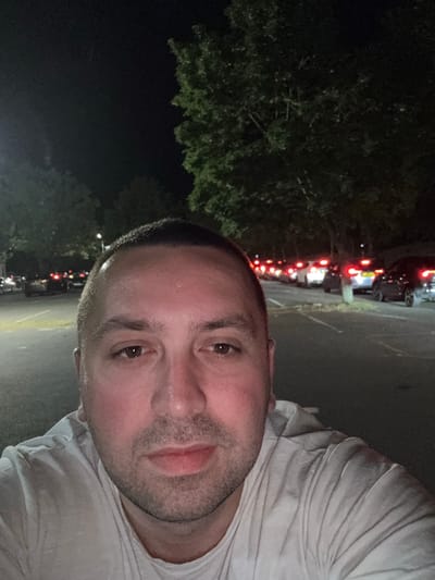 The image is a close-up, selfie-style photograph of a man. He has short, dark hair and stubble, and is wearing a white t-shirt. The background indicates it's nighttime in what appears to be a parking area, based on the presence of vehicles and their lights. A line of vehicles with illuminated tail lights is seen in the background, suggesting a queue or congested traffic. There is also some foliage (trees or shrubs) on the left side of the frame, further indicating an outdoor setting. Based on the location provided, Borough of Spelthorne, United Kingdom, the scene is possibly in a public space with parking facilities.
