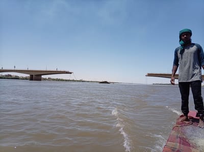 A medium shot shows a man standing on the bow of a boat traveling on the Nile River. The man has dark skin and is wearing a green headscarf, a long-sleeved gray shirt, and dark pants. He is barefoot. The boat is small and has a red and tan patterned cover on the bow. A fishing net lies partially on the cover. The river water is brown and choppy.

In the background, there is a damaged bridge, broken in the middle, spanning the river. Vegetation is visible on the riverbanks. The sky is blue and clear. This scene is in the city of Bahri, Sudan.